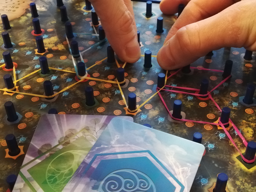 Pour m’étaler et placer un élastique entre 4 picots, je dois défausser 2 cartes vertes et jaunes, puis donner une carte bleue à mon adversaire car il occupe déjà un picot. C’est bien, je vais gagner 5 piépièces, mais que va-t-il faire avec cette carte ? Peut-être pire que moi !